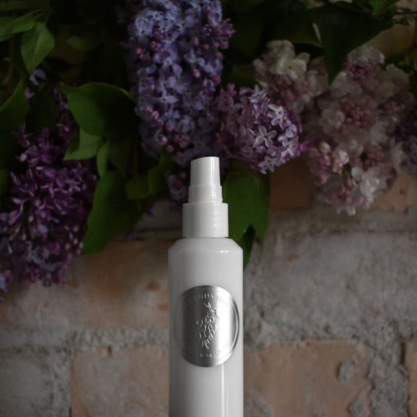 White spray bottle with a silver label on a textured surface with purple flowers in the background. TIDELANDS HOUSE Lilac Nectar Artisanal Copper Distillate