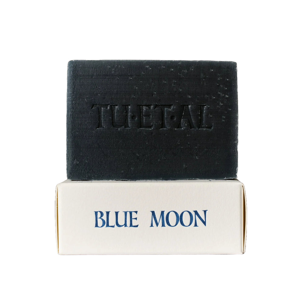 A dark charcoal gray bar of TU·ET·AL Blue Moon soap resting on top of its white cardboard packaging box, which features the scent name 'BLUE MOON' in blue lettering.
