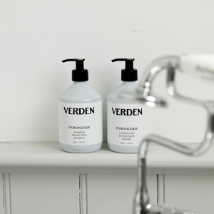 Two white pump bottles of VERDEN haircare products, specifically D'Orangerie Shampoo and Conditioner, sitting on a white shelf next to a blurred chrome faucet.