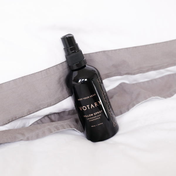 Black bottle of Votary Pillow Spray on a bed with gray pillows.