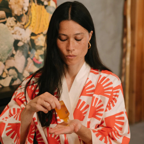 Woman in a red and white robe applying a face oil to her hand with a painting in the background.