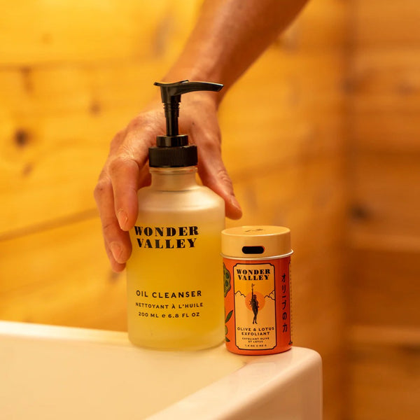 Hand holding a 'Wonder Valley' oil cleanser bottle with olive & lotus exfoliant next to it on a sink.