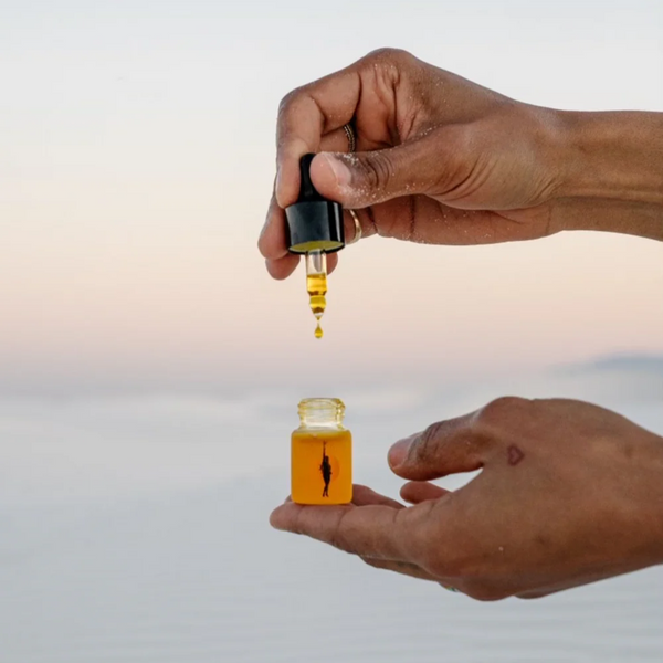 Hand holding a dropper of mini Face Oil by Wonder Valley and in the other hand she is holding the glass bottle of this orange colored oil.
