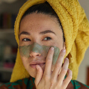 Woman applying green facial mask with yellow towel on head