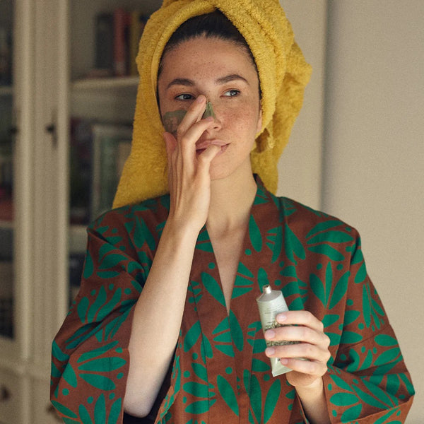 Woman applying Wonder Valley mud mask to her face with a yellow towel on her head, wearing a patterned robe.