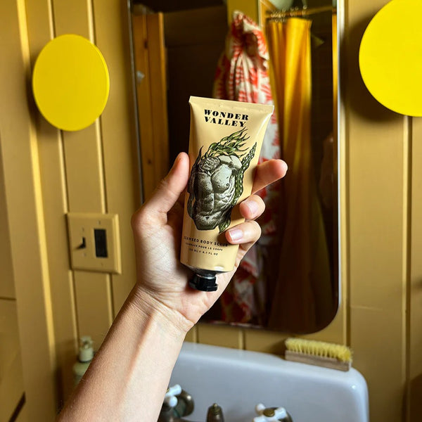 Hand holding a tube of Wonder Valley body scrub in front of a bathroom mirror.