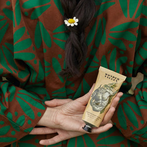 Person holding a tube of Wonder Valley body scrub product with a floral hair accessory, wearing a patterned garment.